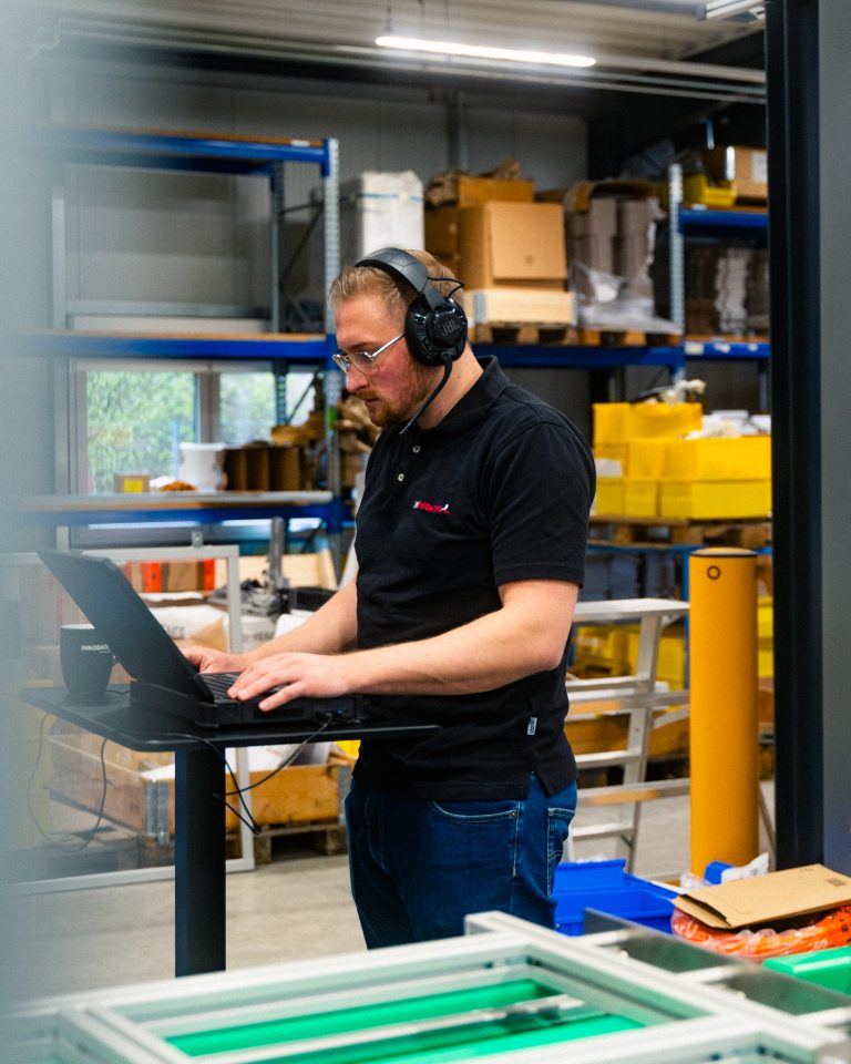 Danny Hapke mit Brille und Kopfhörer steht an einem Stehtisch in einer Industriehalle und arbeitet an einem Laptop. Er trägt ein schwarzes Poloshirt mit dem Logo „INKODATEC“. Im Hintergrund sind Regale mit gelben und braunen Lagerboxen sowie Kartons zu sehen. Die Umgebung wirkt ordentlich und technisch organisiert.